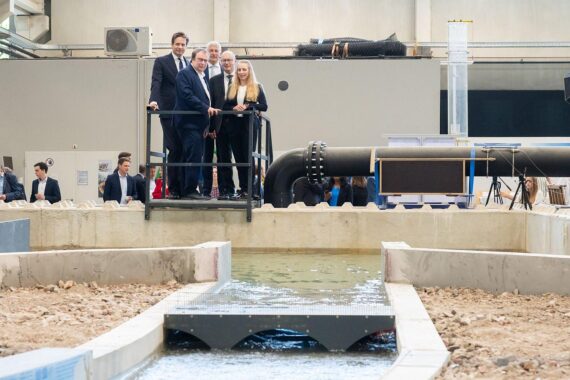 Brückenmodell: Wasser staut sich, Wissenschaftlerin erklärt Hochwasserschutz, Politiker Oliver Krischer und Vincent Karremans lauschen.