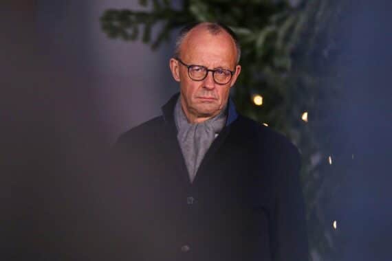 Friedrich Merz beim Weihnachtsbaumschlagen, umgeben von Tannen, mit einem nachdenklichen Gesichtsausdruck.