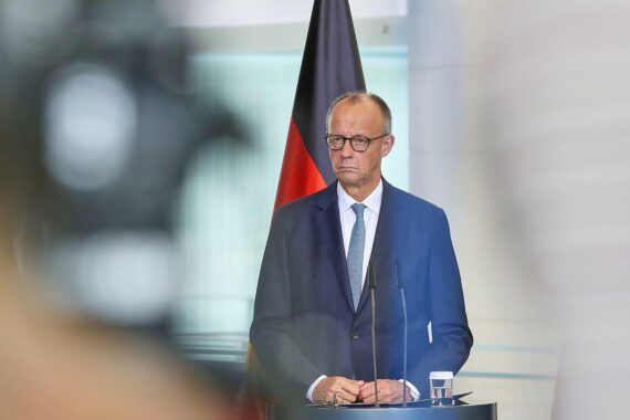 Friedrich Merz an einem Tisch, umgeben von politischen Symbolen, diskutiert Menschenrechte im NATO-Gipfel.