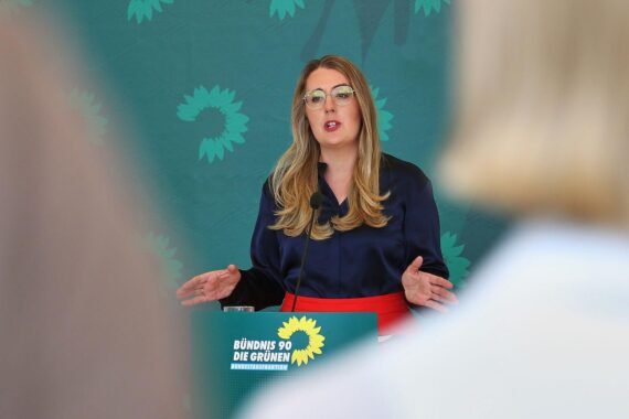 Katharina Dröge vor Hintergrund von Protest und KI-Rechenzentren, warnend vor gesellschaftlichen Konflikten.