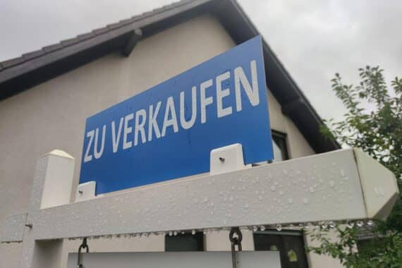 Immobilienmarkt: „Zu verkaufen“-Schild vor Stadtgebäuden, im Hintergrund Wohn- und Gewerbeimmobilienpreise im Trend.