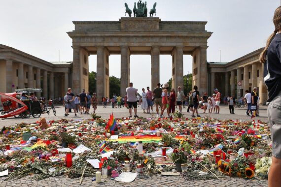 Trauernde Menschen nach CSD-Anschlag in Berlin, 31.07.2026, im Hintergrund Polizisten und Blumen.