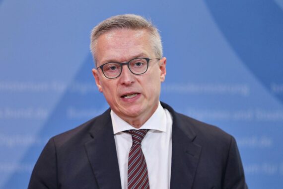 Karsten Wildberger im Archiv, mit KI-/Wachstumssymbolen, die Deutschlands Wirtschaftsboom durch Künstliche Intelligenz andeuten.
