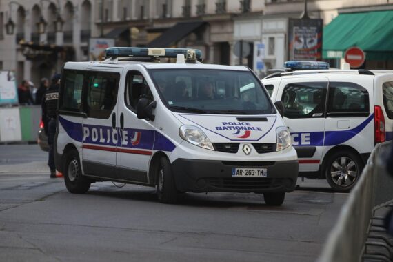 Französisches Polizeiauto (Archiv) vor Hintergrund von Protesten, Gewalt, brennenden Autos und zerstörten Schaufenstern.
