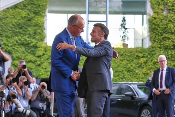 Friedrich Merz und Emmanuel Macron (Archiv) vor dem Hintergrund europäischer Autoindustrie und Verbrenner-Aus.