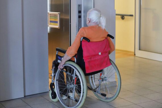 Pflegeheim: Frau sitzt im Rollstuhl, im Hintergrund Angehörigen-Unterstützung in Niedersachsen.