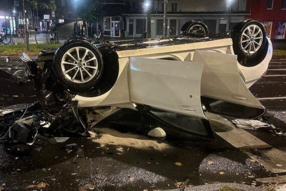 Ein auf dem Dach liegendes Unfallauto neben einem abgeknickten Ampelmast, umgeben von Einsatzkräften.