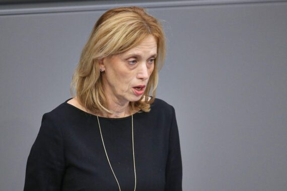Karin Prien bei einer politischen Rede, im Hintergrund symbolische KI-Technologie und Steuerdokumente.