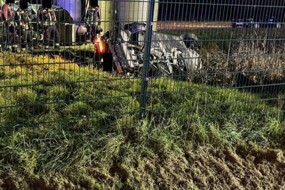 Ein verunfallter BMW liegt neben einem durchbrochenen Zaun auf der linken Fahrzeugseite an einem Regenrückhaltebecken.