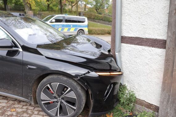 Ein schwarzer Elektro-Sportwagen mit schwer beschädigter Front vor der eingedrückten Wand einer Fachwerkscheune.