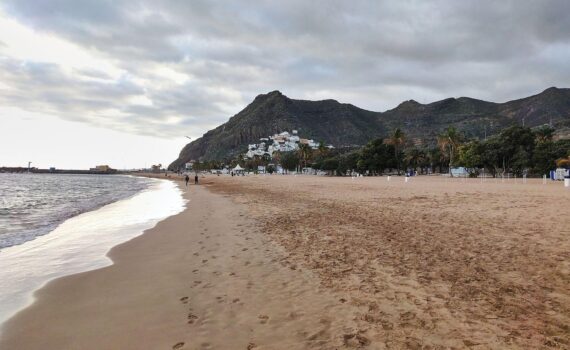 Ein Strand auf Teneriffa mit Urlaubern, Sonnenschirmen und Hotels als Symbol für steigende Reisepreise.