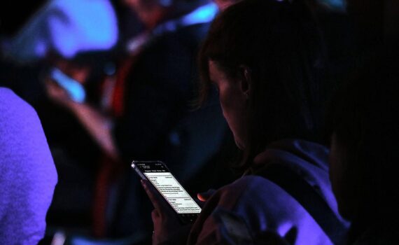 Eine junge Frau blickt unsicher auf eine Finanz-App ihres Smartphones, daneben liegen Rechnungen und Budgetnotizen.