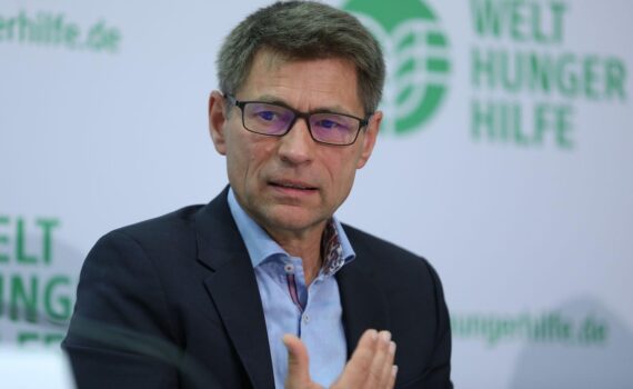 Mathias Mogge (Archiv) vor Bildern zu Gaza-Hilfen: teure Lebensmittel, Nahrungsmittelhilfe und Wasser-Sanitärversorgung.